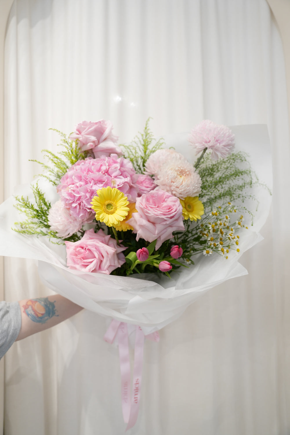 Bouquet of flowers by amicis flower shop held by a person against a white curtain