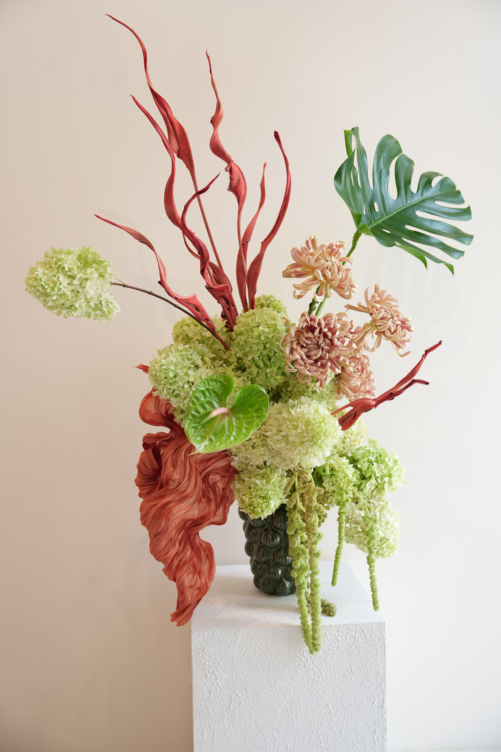 Decorative floral arrangement with red coral-like elements and green leaves on a white pedestal against a beige background. by amicis flower shop