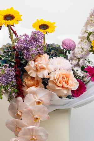 Bouquet of colorful flowers including yellow sunflowers, purple and pink flowers, and white orchids on a white background.