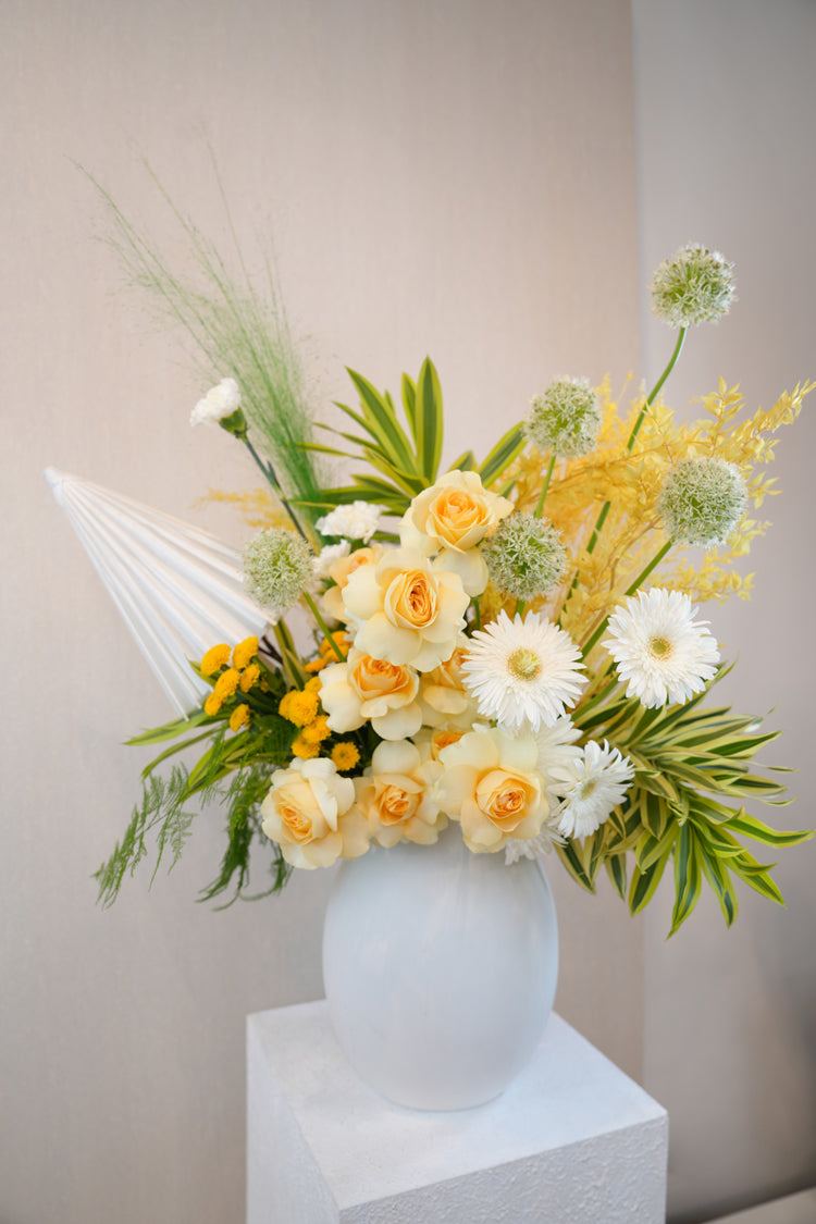 Parasol Twist - cream roses, white gerberas, green alliums, bright yellow chrysanthemums, and a white fan in a white ceramic vase by Amicis