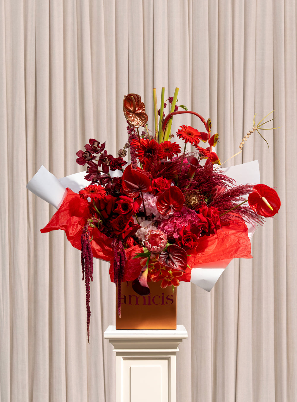 Bordeaux deep red arrangement with crimson roses, red anthurium, burgundy cymbidium orchids and trailing amaranthus in a copper Amicis box