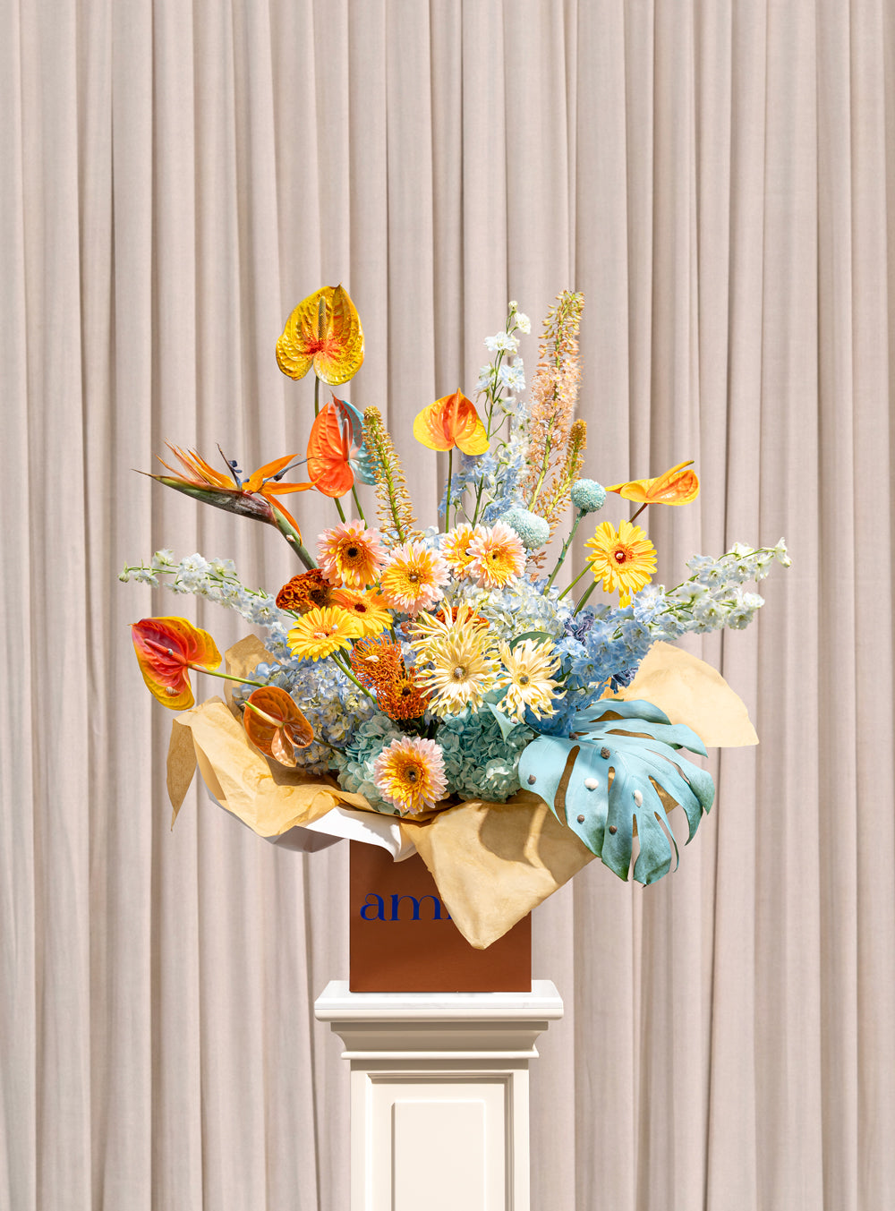 Elysian tall arrangement with yellow and coral anthurium, bird of paradise, blue delphinium and gerbera in a copper Amicis box