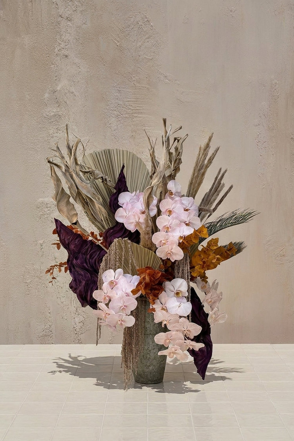 Amaranthine - blush orchids, deep purple leaves, ochre dried elements, pampas grass, and dried palms in a textured grey vase by Amicis