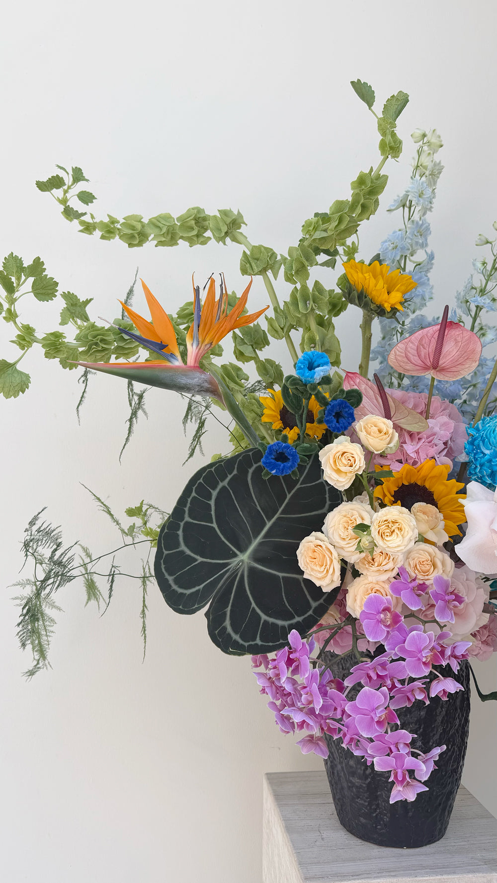 Sweet Love - orange Bird of Paradise, yellow sunflowers, blue delphinium, lavender orchids, and mixed roses in a dark textured vase by Amicis.