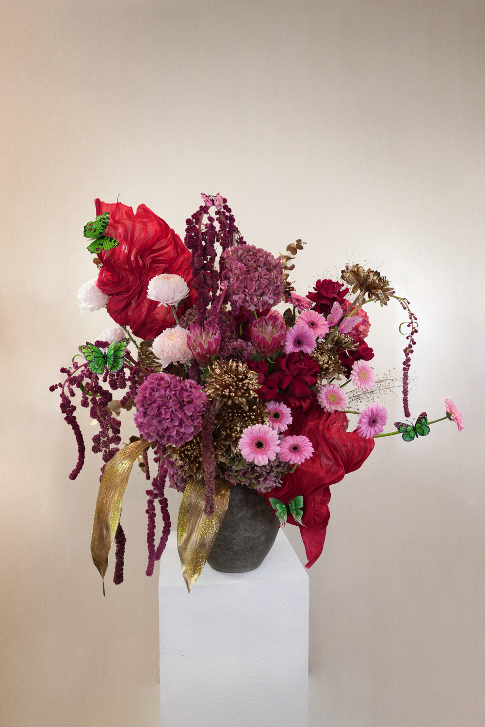 Decorative floral arrangement with red coral-like elements and green leaves on a white pedestal against a beige background. by amicis flower shop