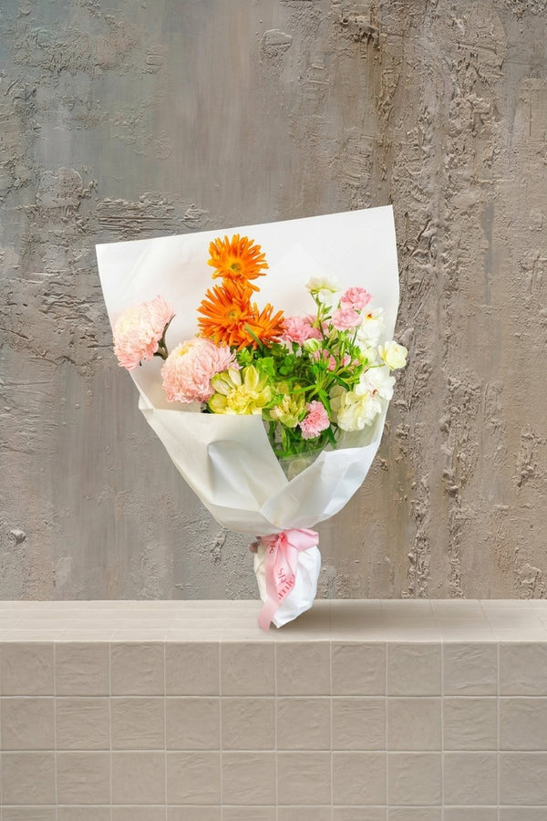 Sunset Whimsy - orange gerberas, blush pink carnations, and cream spray flowers in a white wrapped bouquet with pink ribbon by Amicis