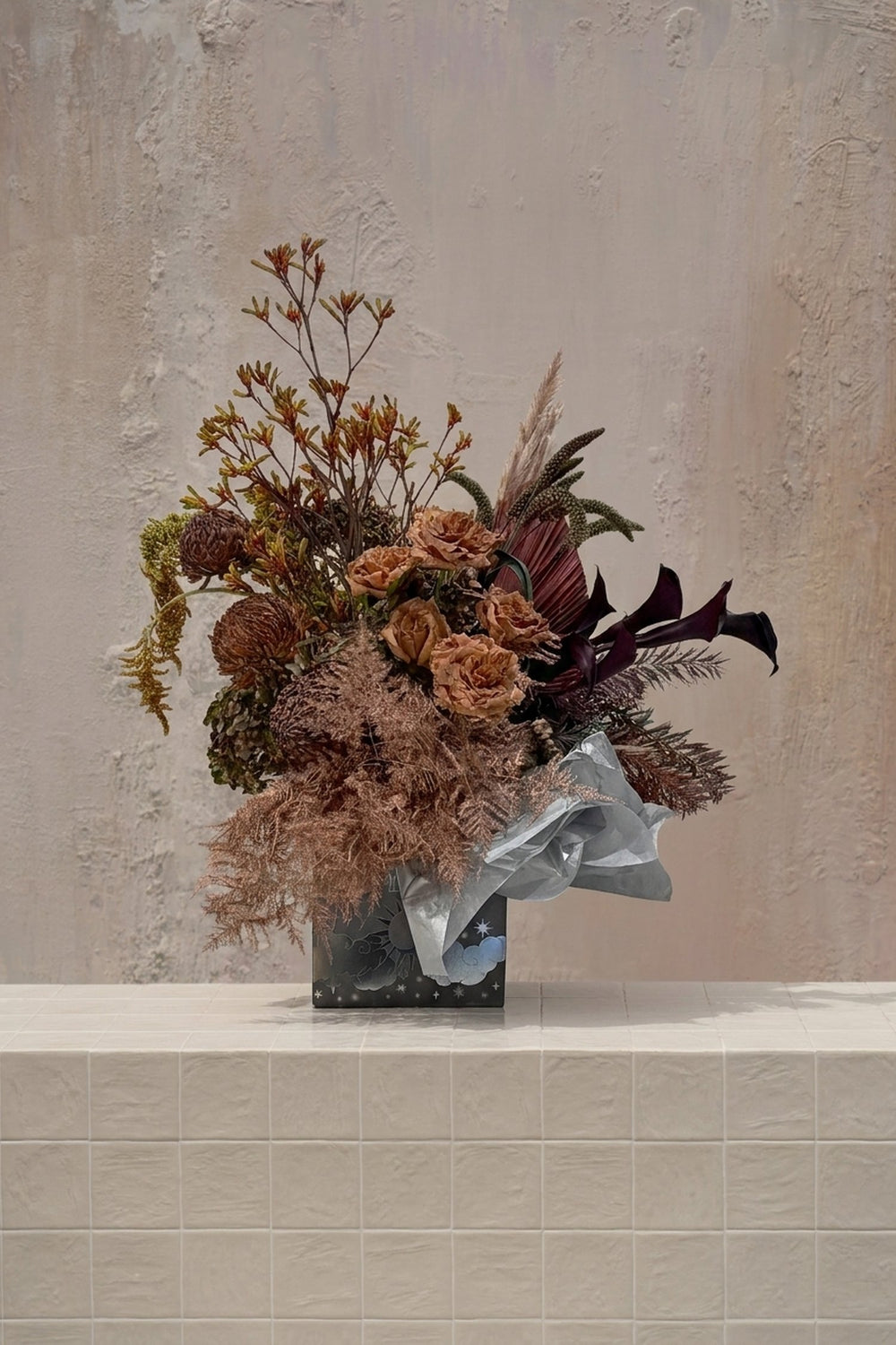 Eclipse 12 - burnt orange roses, dark calla lilies, and brown protea with dried grasses in a dark decorative box with blue tissue by Amicis