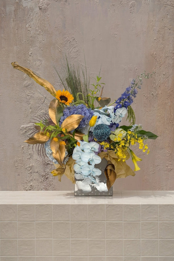 Eclipse 13 - vibrant blue hydrangeas, light blue orchids, yellow sunflower, delphinium, and golden foliage in a silver embossed vase by Amicis