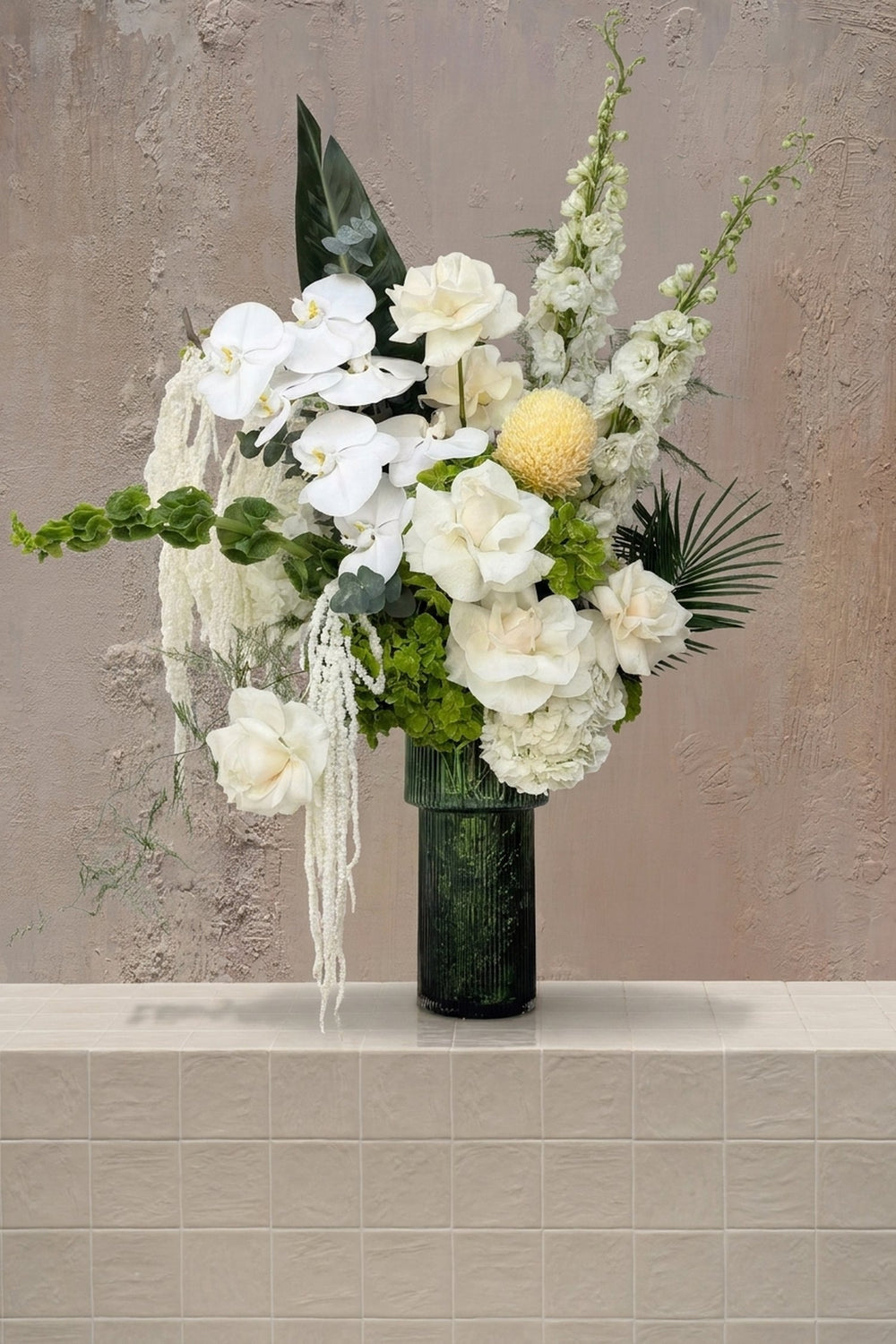 Garden Kiss - white orchids, cream roses, white delphinium, and trailing white and green amaranthus in a dark green ribbed glass vase by Amicis.