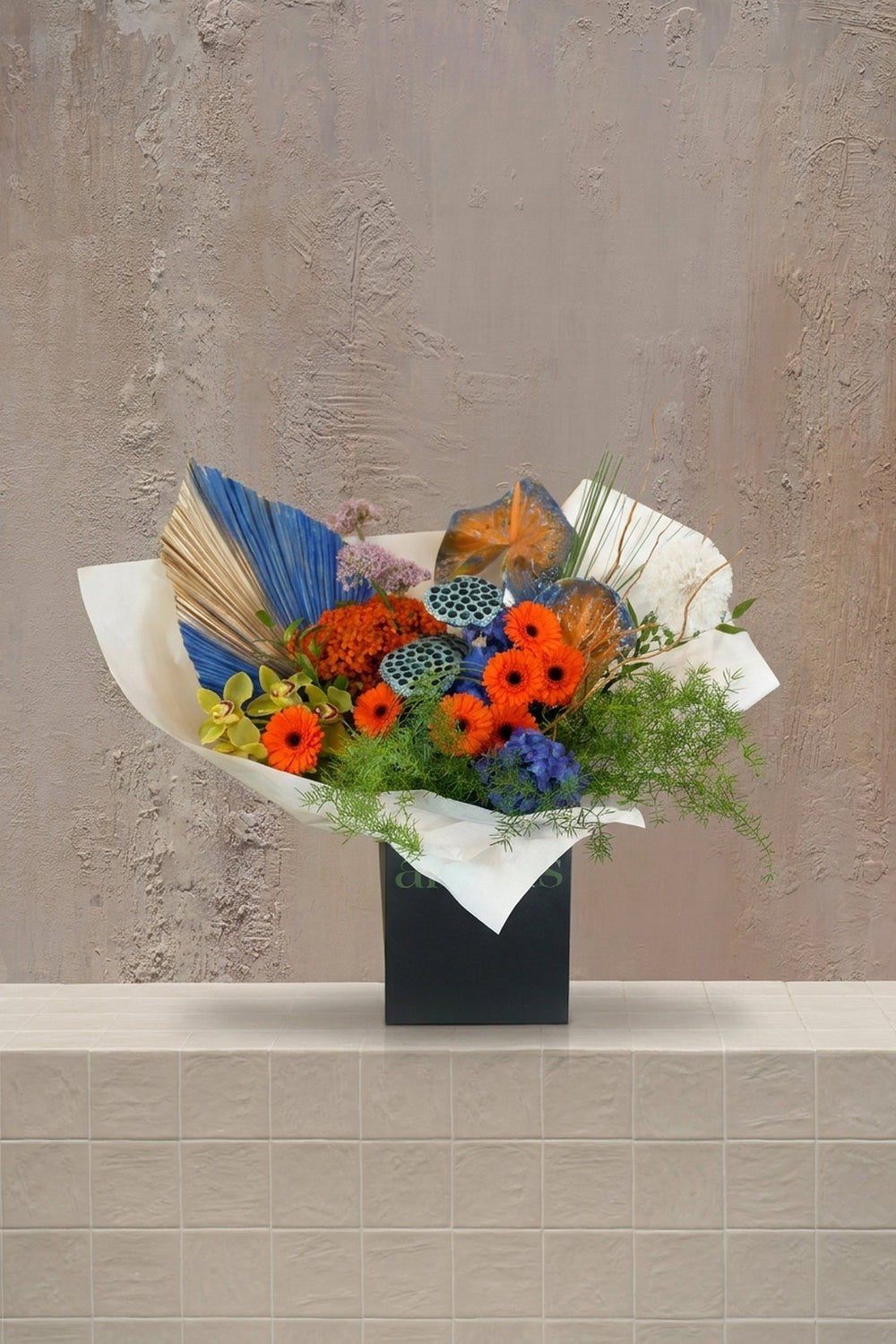 Ember Sonata - vibrant orange gerberas, green cymbidium orchids, blue delphinium, and blue painted palm fronds in a black gift box by Amicis