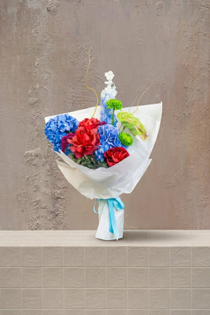 Prism Bloom - deep blue hydrangeas, red roses, green chrysanthemums, anthurium, and white delphinium in a white wrapped bouquet by Amicis