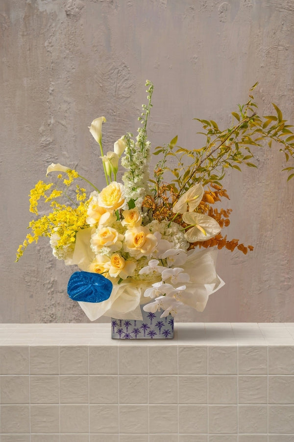 Hilal Bloom - yellow roses, white orchids, calla lilies, cream anthurium, and delphinium in a blue palm-patterned box wrapped in tissue by Amicis.