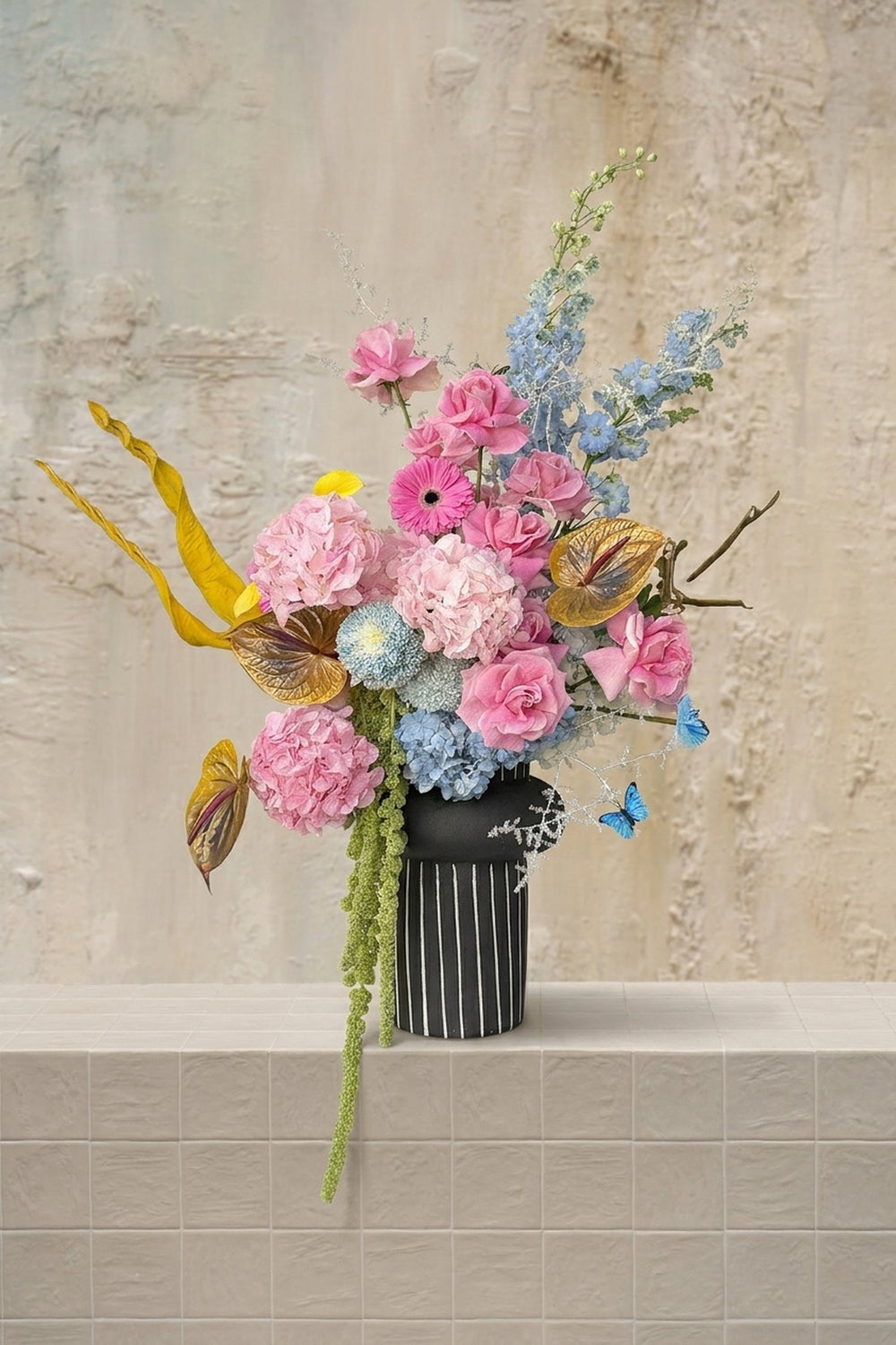 Pastel Fairy - blush pink roses, hydrangeas, light blue delphinium, gold anthurium, and trailing amaranthus in a black striped ceramic vase by Amicis.