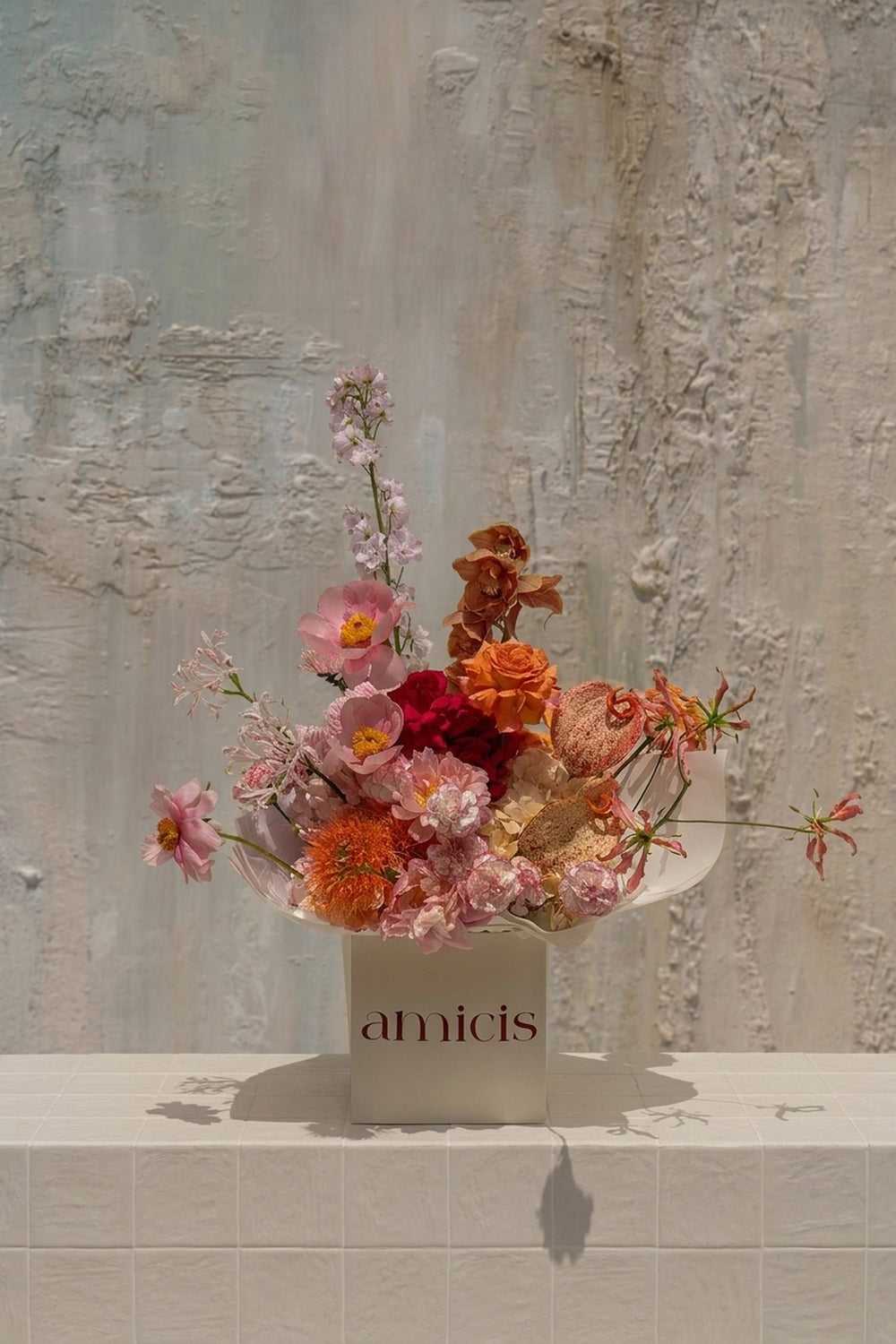 Radiant Symphony - blush pink poppies, orange roses, red anthurium, pincushion protea, and gloriosa lilies in a sculptural white vase by Amicis
