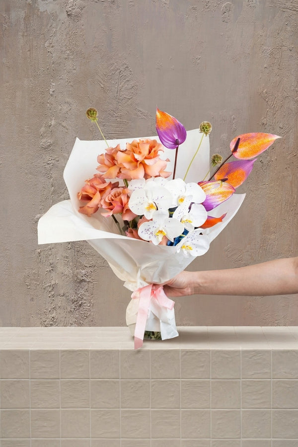 Orchid Mirage - white orchids, peach roses, purple-orange anthurium, and green alliums in a white wrapped bouquet with pink ribbon by Amicis