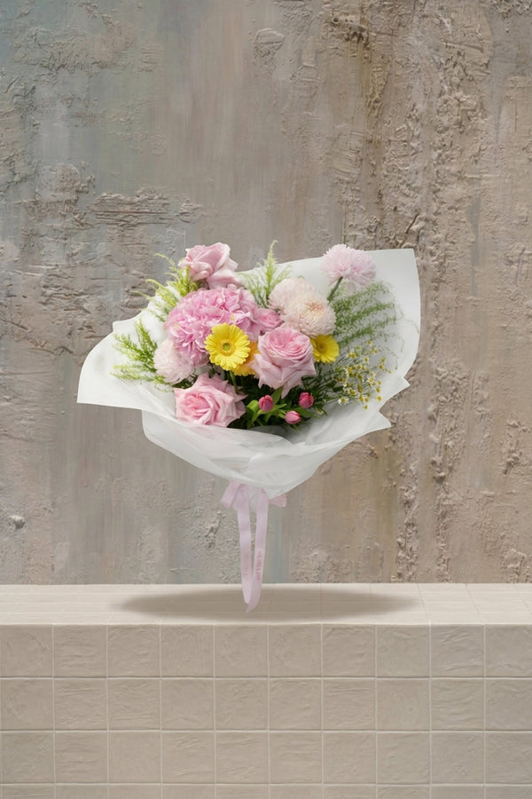 Evangeline - blush pink roses, pink carnations, yellow gerbera, white mums, and pink tulip buds with greenery in a white wrapped bouquet by Amicis.