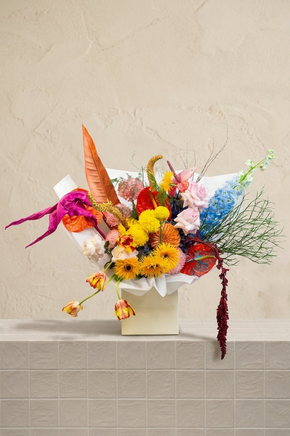 Spring Blossoms - red anthuriums, yellow gerberas, blush roses, blue delphinium, orange tulips, and trailing amaranthus in a white box by Amicis.
