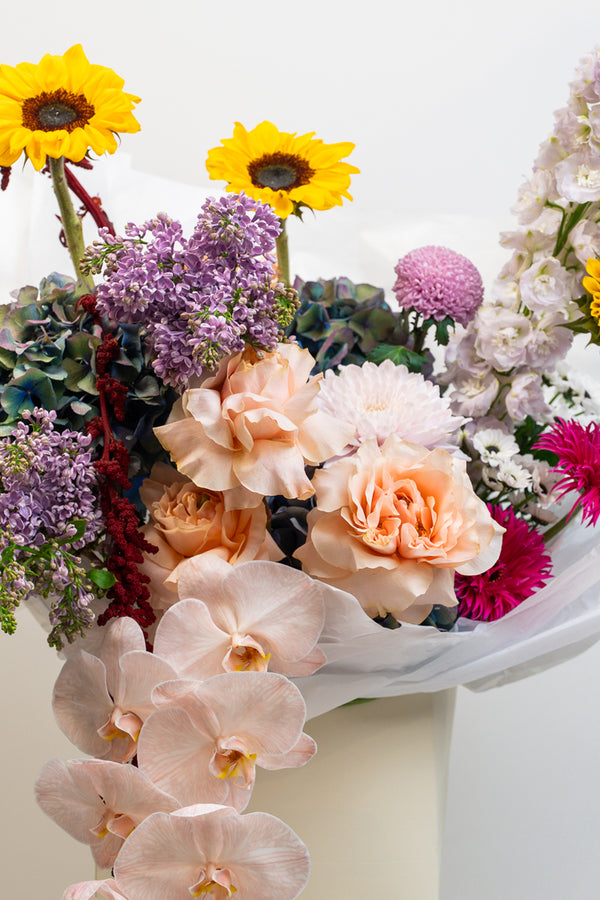 Bouquet of colorful flowers including yellow sunflowers, purple and pink flowers, and white orchids on a white background.