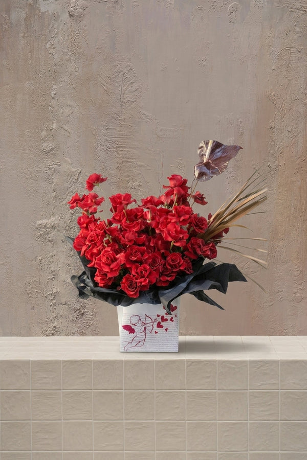 The Love Edit - deep red roses, dark burgundy anthurium, and golden grasses in a white box with cupid drawing and black wrap by Amicis