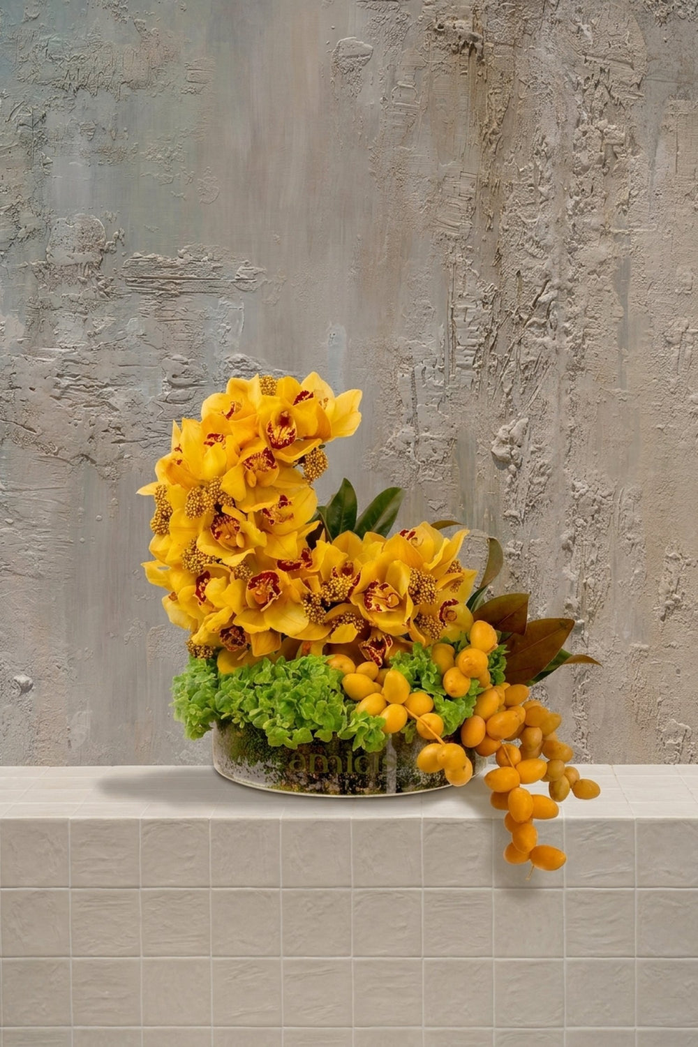 Twilight Bloom - yellow Cymbidium orchids with red centers, green foliage, and trailing orange dates in a textured ceramic bowl by Amicis